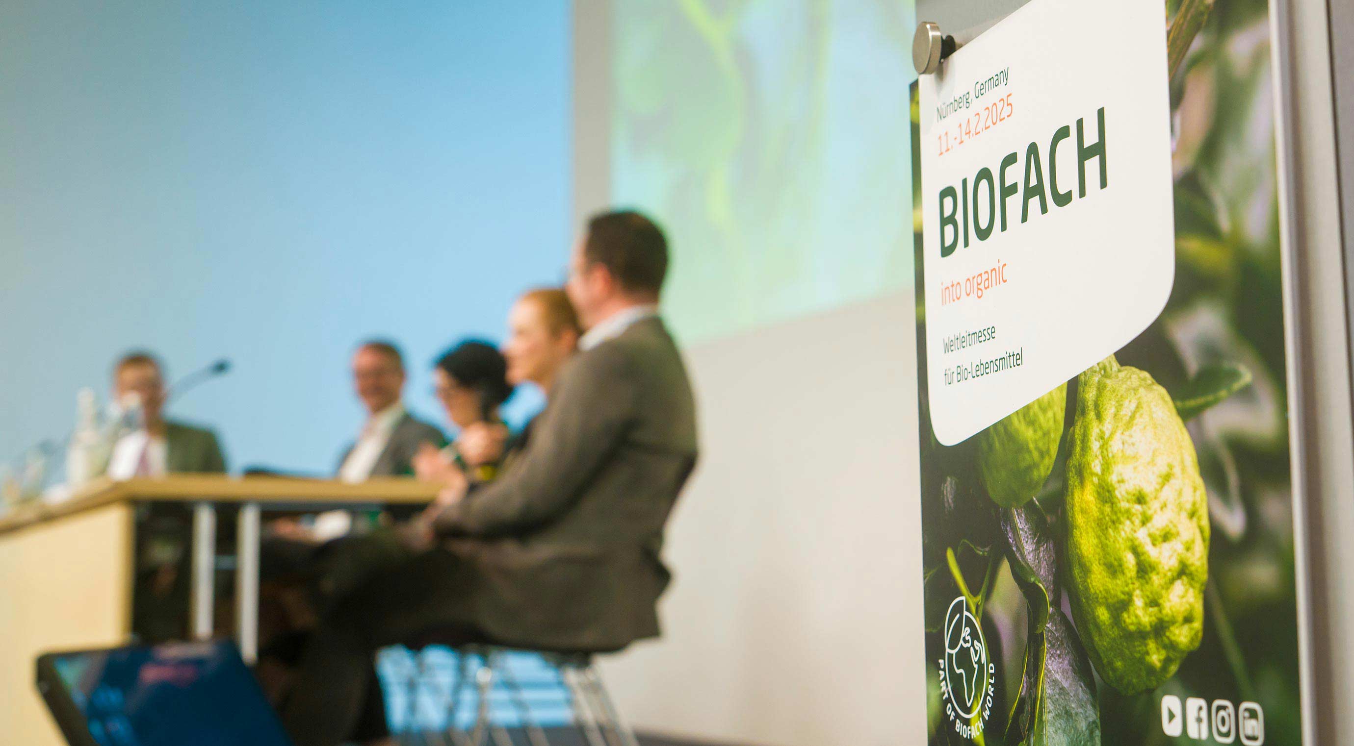 Close-up of a BIOFACH sign. In the background, some speakers can be seen out of focus at a table on the stage.