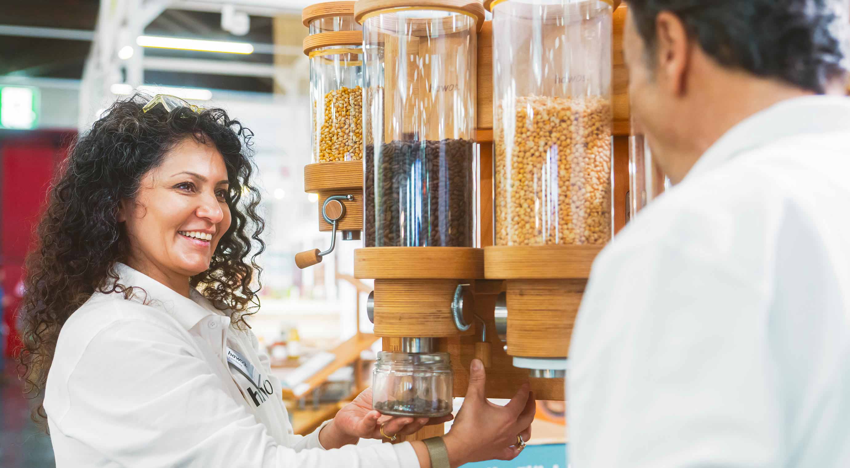 An exhibitor drops coffee beans from a container into a storage jar and looks at a visitor